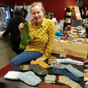 Photo of Anna Makila sitting on the edge of a table which has lots of pairs of knitted socks on it. She is smiling to camera and holding up a pair of socks.