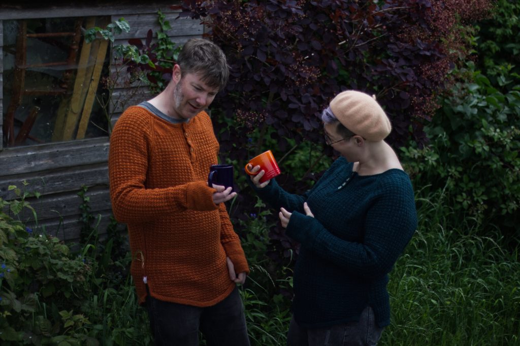 two people wearing knitted henley jumpers, one orange and one deep dark green. they are holding mugs in similar colours but opposite to the jumpers they are wearing. 